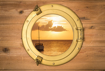 Vintage brass porthole in wooden wall with view to a boat in sunset