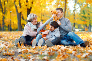 Happy family sitting on fallen leaves, playing and having fun in autumn city park. Children and parents together having a nice day. Bright sunlight and yellow leaves on trees, fall season.