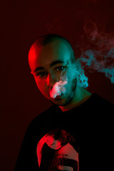 Close-up studio shot of a young bald guy vaping, blowing out a cloud of smoke on red background.