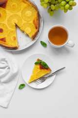 Sliced upside down pineapple cake decorated green leaves with cup of tea on white table. Summer tropical dessert. Top view. Copy space