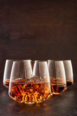 A crystal glass of whiskey on the rocks with a double portion on a wooden table. In the background, three glasses with one shot of whiskey.
