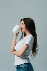 smiling girl with closed eyes in white t-shirt drinking from white mug isolated on grey
