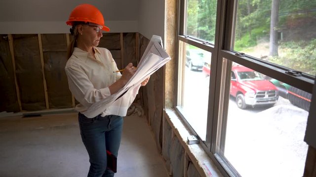 Female construction project manager with hard hat doing walkthrough inspection of new house construction with blueprints.