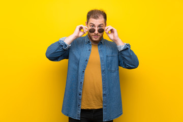 Blonde man over isolated yellow wall with glasses and surprised