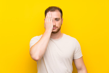 Handsome man over yellow background covering a eye by hand