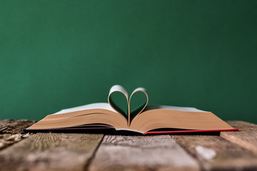 Back to school concept, open book and pages folded in the shape of a heart on an old wooden surface on a backdrop of clean green chalk board, selective focus, copy space