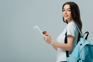 side view of pretty smiling girl with blue backpack using digital tablet isolated on grey