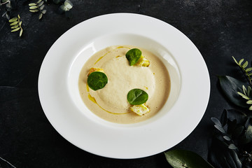 Exquisite Serving White Restaurant Plate of Cream Cappuccino Soup with Porcini Mushrooms and Truffle Close Up. Beautiful Creative Molecular Dish with Boletus on Black Stone and Plants Background
