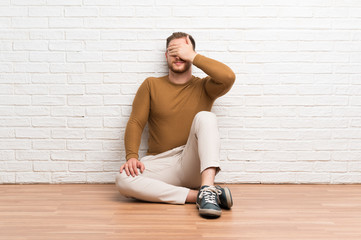 Blonde man sitting on the floor covering eyes by hands
