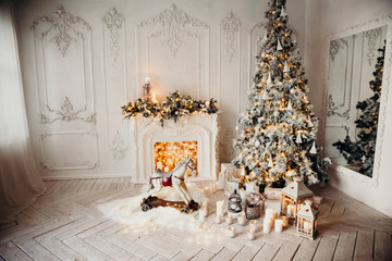 classical luxury interior of a white room with Christmas tree with garland, decorated fireplace, rocking horse, gifts for new year