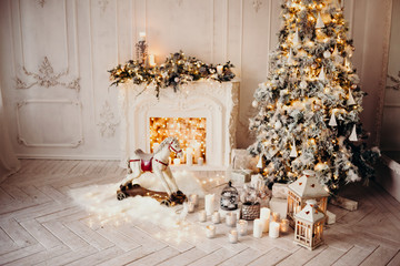 classical luxury interior of a white room with Christmas tree with garland, decorated fireplace, rocking horse, gifts for new year