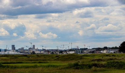 Fototapeta na wymiar Cranes along the Dublin skyline with the green grass of Bull Island causeway in the foreground and blue sky and white fluffy clouds above the city scene.