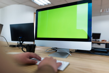 Hombre frente a pantalla de computador escribiendo en pantalla verde, con teclado y mouse junto a un café y más pantallas