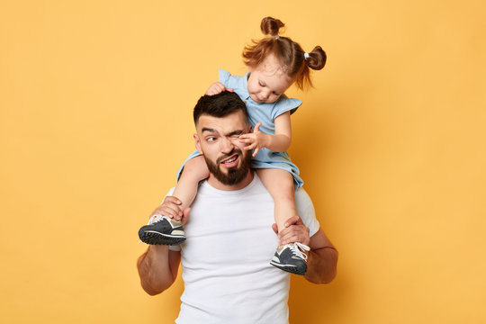 naughty behavior. parent cannot handle with kid with much energy. girl tiuching her dad's eyes while sittting on shoulders . close up photo. isolated yellow background. unacceptable kind of behaviour