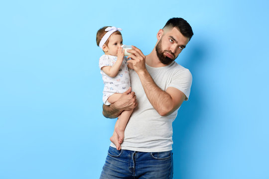 exhausted , tired father feeding his baby girl with milk bottle. close up photo. isolated blue background. studio shot. poor dad is fed up loking after child.
