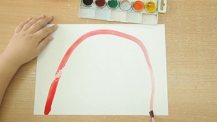 The child holds a brush in his right hand and draws a rainbow on a white sheet of paper. Red line of paint on a sheet of white paper