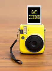 Bright yellow instant camera with photo that reads "say cheese".  Wooden background. Shallow depth of field.