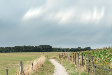 Dirt pathway between fence and corn field under cloudy sky. Long exposure shot.