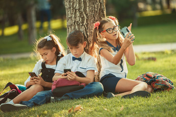 Three school friends are sitting on the grass under a tree and playing their gadgets. Friends do not communicate in life preferring the phone