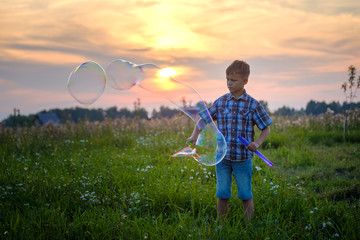cute boy blowing big soap bubbles in the meadow in summer on the background of a beautiful sunset