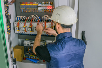 An electrician in a helmet connects the wires of the circuit breakers in the electrical box. A...