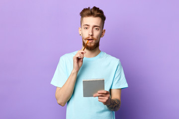 thoughtful serious man holding pen near lips and notepad isolated on blue background. close up portrait. free time, spare time, lifestyle. guy making up a story