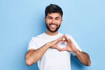 handsome cheerful guy is making a heart shape symbol with his fingers, expresses love and positive romantic feeling. Isolated on a blue background, positive man - 284005436