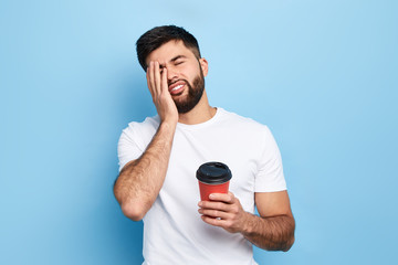 Tired exhausted man covers face with palm, holds disposable paper cup of coffee. man doesn't want...