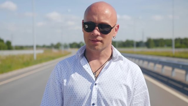 Portrait of a bald man in sunglasses singing and walking along the road. Medium shot.