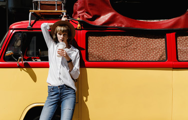 Stylish young woman in a straw hat dressed in white shirt and jeans drinking a milkshake standing...