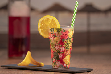 Slice of orange and a bottle of cocktails behind a tropical juice served with a straw