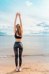 Slender athletic girl in sports clothes doing exercise on the beach against the beautiful sky