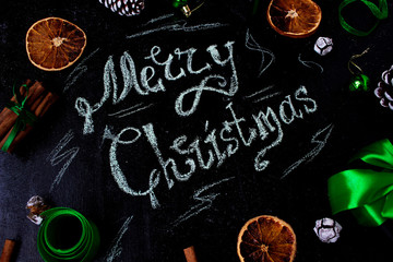 Christmas background with a words:Merry christmas, dry orange,white pinecone,green christmas tree balls,ginger sticks,green ribbon on a black background,flat lay