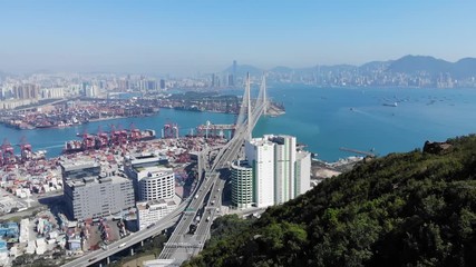 Woman stay on rock at top, stretch up hands, camera fly over. Hong Kong city panorama and large Stonecutters bridge seen ahead below. Hiker girl enjoy beautiful scenery from viewpoint at peak of mount