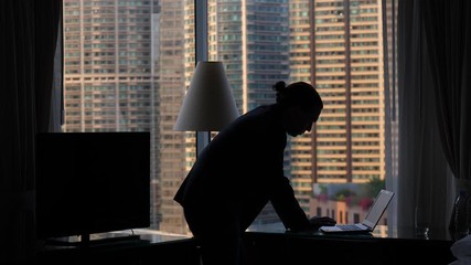 Man stay bend over laptop, silhouetted shot against room window. Dim natural light at apartments, guy work or check something online, type text using portable computer keyboard