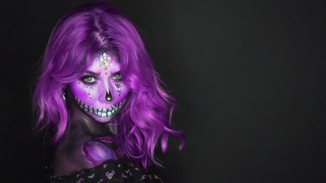 A young woman looks around sharply waving purple hair. Beauty shows creative makeup sugar skull. Greasepaint with glitter, sparkles, stones, body art skeleton made with paints. Free space for text.