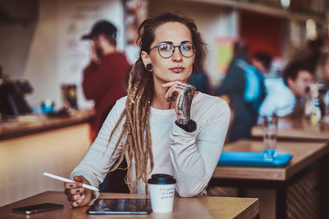 Styilish pensive girl is sketching on digital notepad while sitting at cafe with cup of coffee.