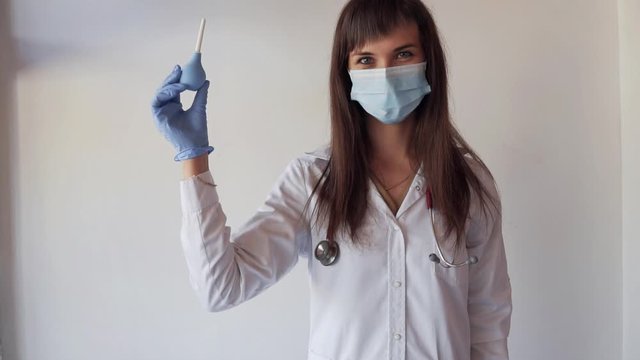 Young nurse in medical mask with stethoscope showing enema on white background