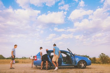 Friends examining broken down car on sunny day at roadtrip vacation excursion. Group of young people on road trip repair breakdown rent car on sea beach. Adventure lifestyle travel friendship concept.