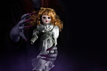 Hands of a witch with long sharp claws holding a dirty porcelain doll with pale face and blind eyes. Halloween, black magic concept.
