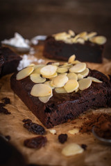 Brownie cake with almond slices placed on paper there are spoon, coco, currant  and flour placed around.