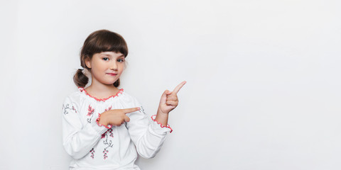 A little Ukrainian girl in a white patterned embroidery on the background of the wall shows with index fingers towards empty space. An emotional little and patriotic child. Creative photography. Copy 