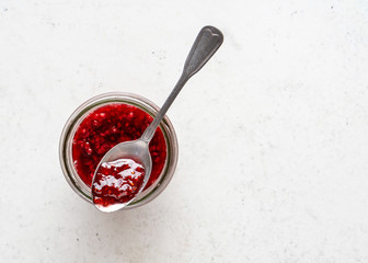 Glass jar of homemade raspberry jam on white stone background.