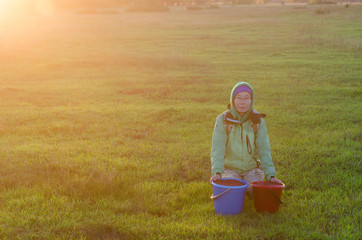 Yakut Asian girl in a hood and hat, and glasses sitting on a green field in the sunset about two...