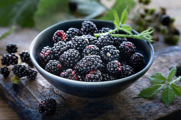 Ripe blackberries are harvested in a ceramic cup. Green leaves around and scattered berries. Wood background
