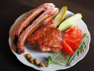 Beautiful smoked pork ribs, tomatoes, salted cucumbers, spicy herbs and mustard in a white plate on a dark wooden table. Appetizing and hearty meat dish with vegetables close-up.