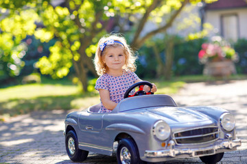 Little adorable toddler girl driving big vintage toy car and having fun with playing outdoors. Gorgeous happy healthy child enjoying warm summer day. Smiling stunning kid playing in domestic garden