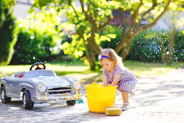 Cute gorgeous toddler girl washing big old toy car in summer garden, outdoors. Happy healthy little...