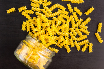 Glass jar filled with dry pasta spiral fusilli and scattered around on black background