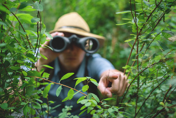 Traveler in hat is looking through binoculars over green forest.